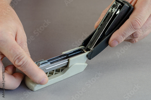 Two Hands holding a stapler against a gray background. A man has opened the stapler device. Part of a series. Close-up. Selective focus.