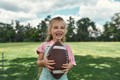 Portrait of cheerful little girl holding an oval brown leather rugby ball and smiling while playing with her parents in park