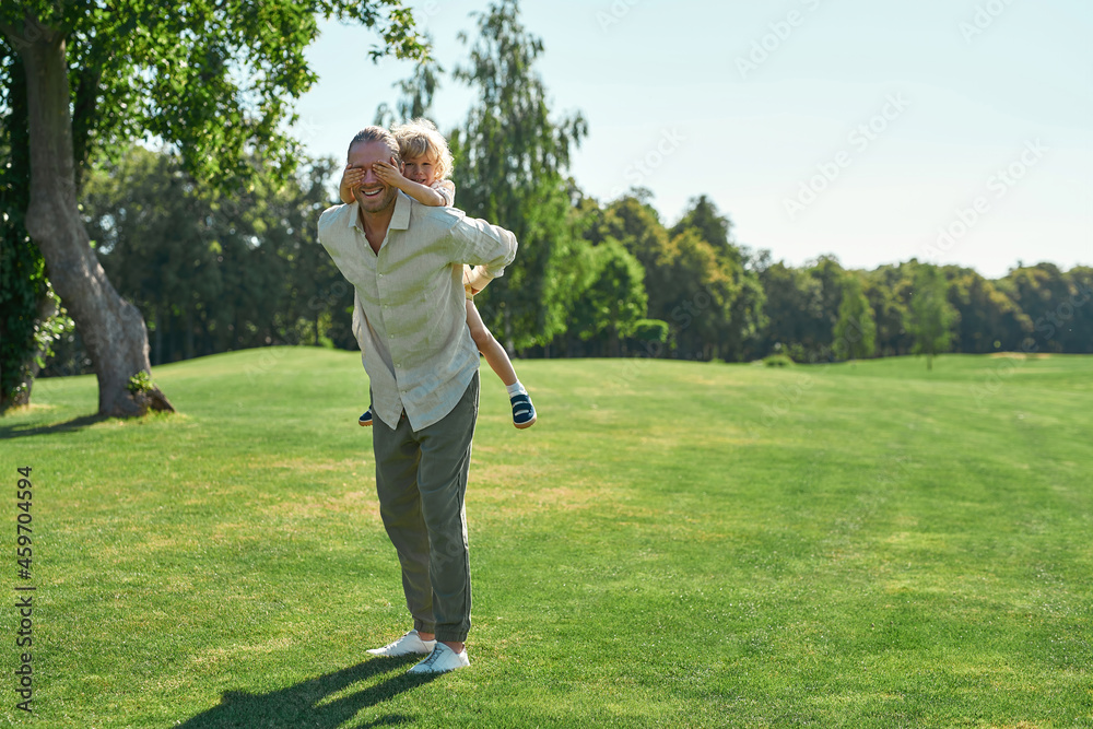 Full length shot of smiling father playing in the park with his little son. Kid having fun, closing dad eyes while sitting on his back Full length shot of smiling father playing in the park with his little son. Kid having fun, closing dad eyes while sitting on his back