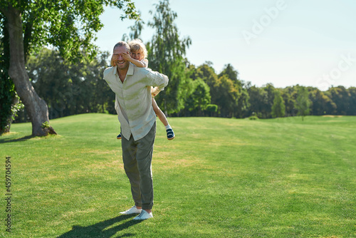 Full length shot of smiling father playing in the park with his little son. Kid having fun, closing dad eyes while sitting on his back