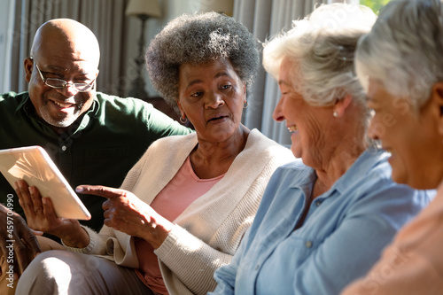 Three happy diverse senior woman and african american male friend sitting on sofa and using tablet
