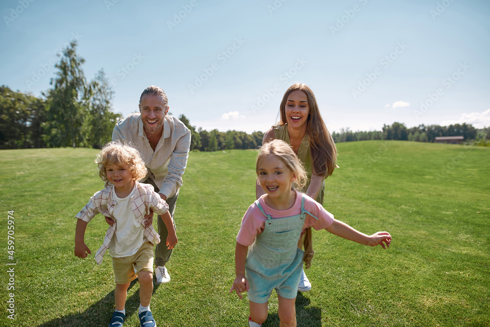 Fototapeta premium Joyful young parents playing catch together with two little children, boy and girl in green park on a summer day