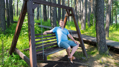 Portrait of an adult man resting in park on a wooden swing on a summer day.