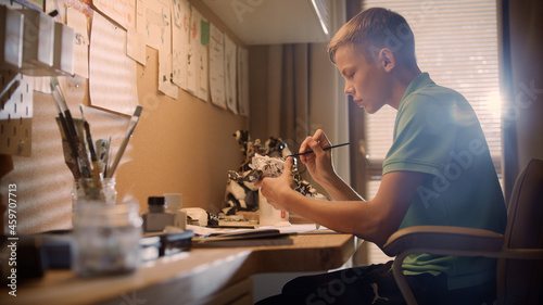 Teenage boy painting a robot skeleton. Focused on work, sitting by a desk in his room