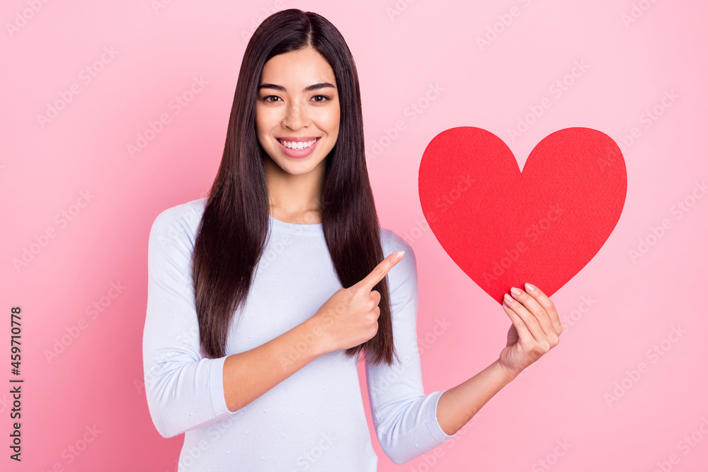 Photo portrait of girl showing finger recommending red heart fell in love isolated on pastel pink color background Photo portrait of girl showing finger recommending red heart fell in love isolated on pastel pink color background