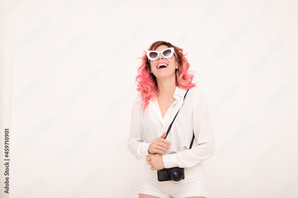 Cheerful woman in sunglasses standing with photo camera against light wall