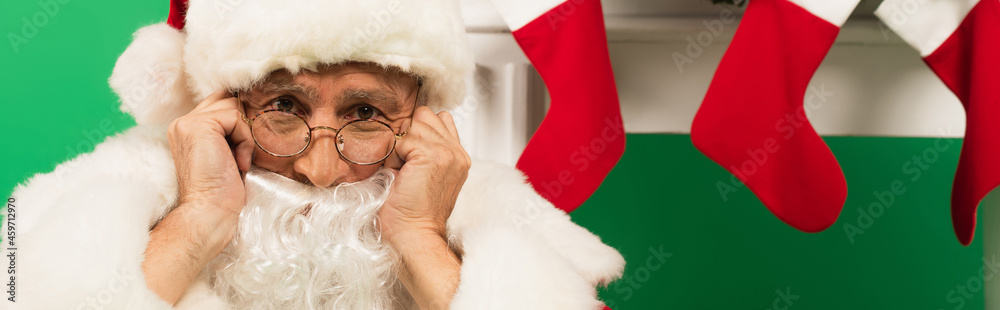 Obraz premium Sad santa claus looking at camera near christmas stockings on fireplace on green background, banner
