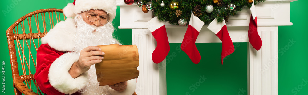 Obraz premium Santa claus holding paper on rocking chair near fireplace with christmas stockings on green background, banner