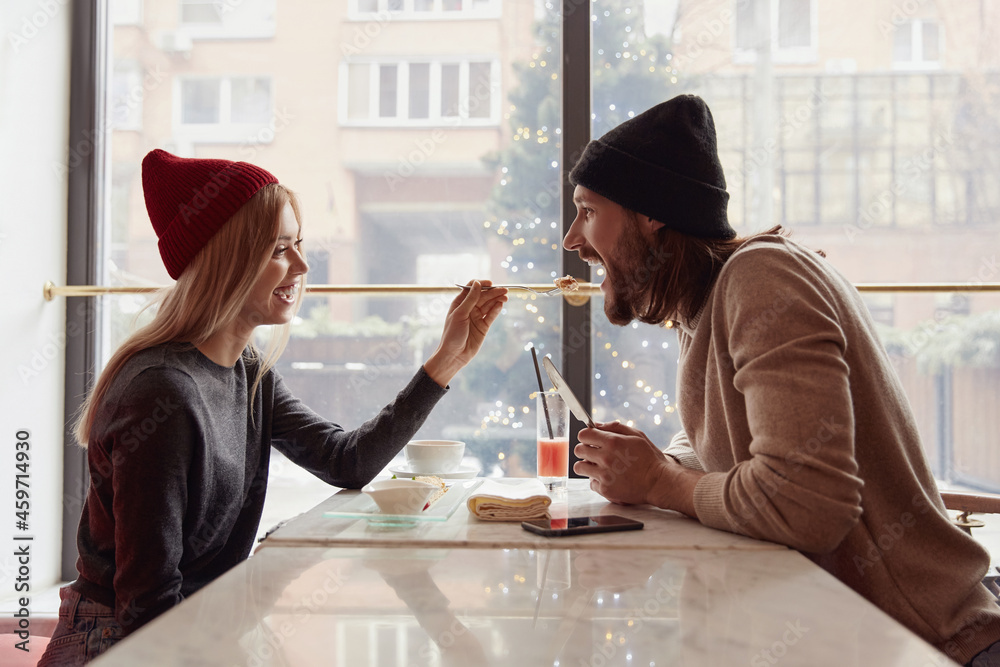 Blonde girl feeding her bearded boyfriend in cafe - obrazy, fototapety, plakaty 