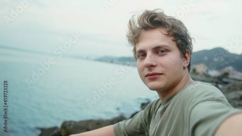 Young blond handsome boy making video call or vlogging on sea and cloudy background he pointing to the sea and smiling
