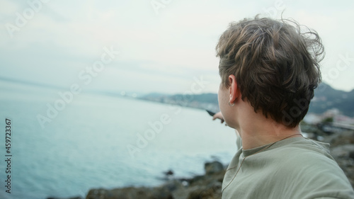 Young blond handsome boy making video call or vlogging on sea and cloudy background he pointing to the sea and smiling