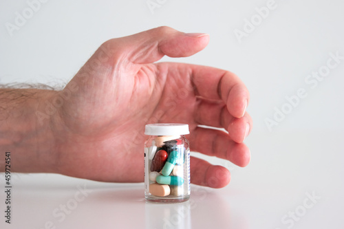 Caucasian male hand reaching out for a glass bottle full of various colored medical pills, tablets and capsules. Close up studio shot, isolated on white background