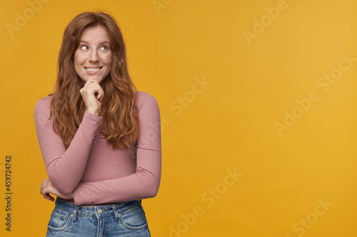 Indoor shot of young ginger woman, with wavy hair and freckles, looks aside at copy space, have dreams or thoughts, isolated.