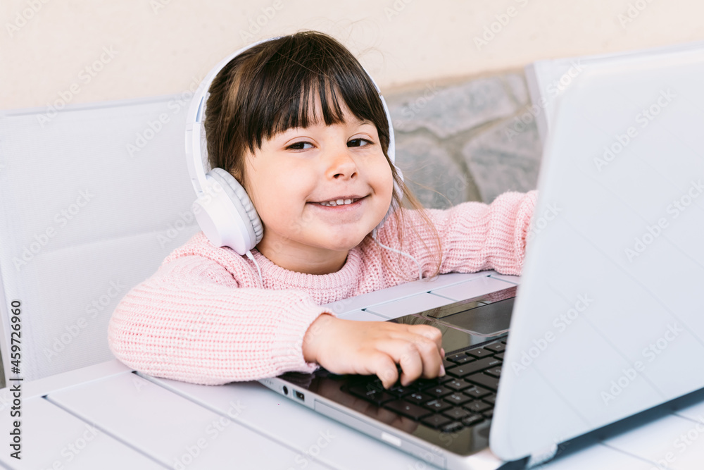 Small girl with white headphones, wearing pink sweater, sitting in front of laptop on terrace, smiling. Concept of distance studying online, childhood and technology. Small girl with white headphones, wearing pink sweater, sitting in front of laptop on terrace, smiling. Concept of distance studying online, childhood and technology.