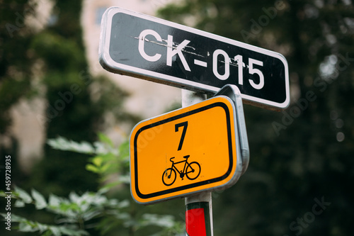 A road sign and a bicycle designation on a background of foliage. Two road signs. One yellow one with a black bike. The second one is black with white lettering