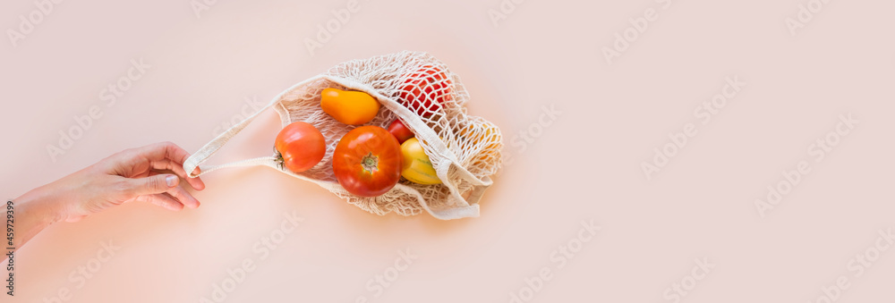 A human hand holds a string bag with tomatoes on a beige background. A human hand holds a string bag with tomatoes on a beige background.