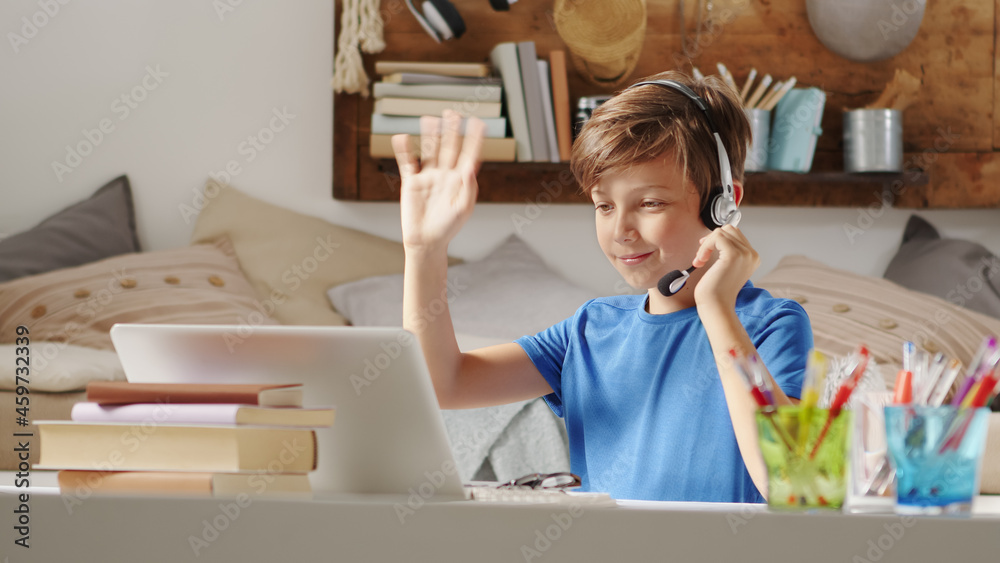 smiling student boy studying at home with remote school connected to computer on the web, raise his hand to answer questions or greet his classmates, sitting at desk full of books, wearing headphones