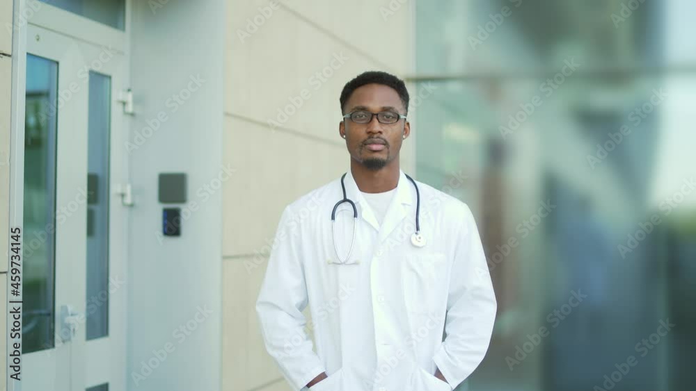 Close up portrait African American doctor looking at camera with arms crossed background of modern hospital outside. Confident happy medic outdoors. male scientist therapist wearing scrubs near clinic Close up portrait African American doctor looking at camera with arms crossed background of modern hospital outside. Confident happy medic outdoors. male scientist therapist wearing scrubs near clinic