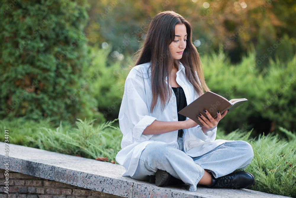 A young and beautiful Arab or Caucasian girl dressed in casual style is reading a book while sitting in the park. Knowledge and education concept. A young and beautiful Arab or Caucasian girl dressed in casual style is reading a book while sitting in the park. Knowledge and education concept.
