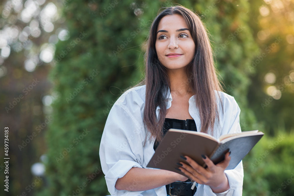 A young and beautiful Arab or Caucasian girl dressed in casual style is reading a book while sitting in the park. Knowledge and education concept.