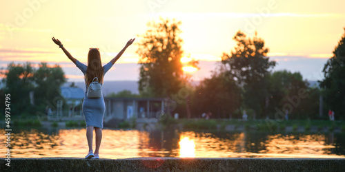 Young woman in casual outfit relaxing on lake side with raised hands on warm evening. Success and wellness concept.