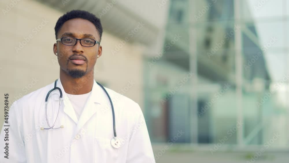 Close up portrait African American doctor looking at camera with arms crossed background of modern hospital outside. Confident happy medic outdoors. male scientist therapist wearing scrubs near clinic Close up portrait African American doctor looking at camera with arms crossed background of modern hospital outside. Confident happy medic outdoors. male scientist therapist wearing scrubs near clinic