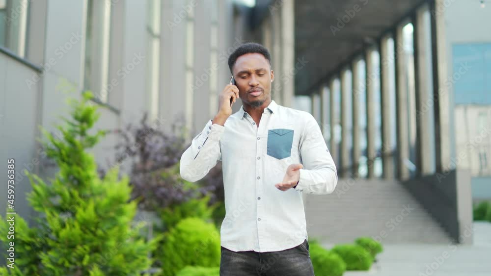 cheerful african american guy walks and talks urban city street in casual clothes using mobile phone Male hipster with smartphone go against modern background Happy smiling male black man conversation