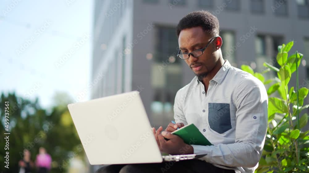 Young african american male student sitting in a city park on a bench with laptop and notebook studying online outdoors Man freelancer in glasses with books learn working remotely in street e-learning