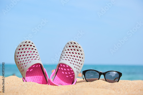 Closeup of clogs shoes and black protective sunglasses on sandy beach at tropical seaside on warm sunny day. Summer vacation concept.