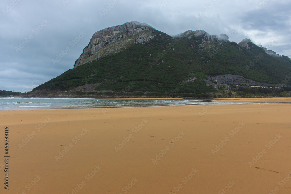 Wide surf beach with mountain in clouds. Autumn landscape with empty coastline. Turquoise seascape. Camino de Santiago landscape. Tourism in Spain. Travel inspiration concept. Beauty in coastal nature Wide surf beach with mountain in clouds. Autumn landscape with empty coastline. Turquoise seascape. Camino de Santiago landscape. Tourism in Spain. Travel inspiration concept. Beauty in coastal nature