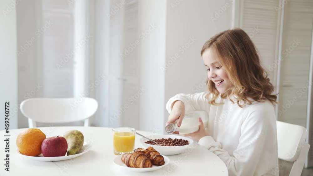 A side view of the happy little girl pouring milk over the cereal at breakfast A side view of the happy little girl pouring milk over the cereal at breakfast