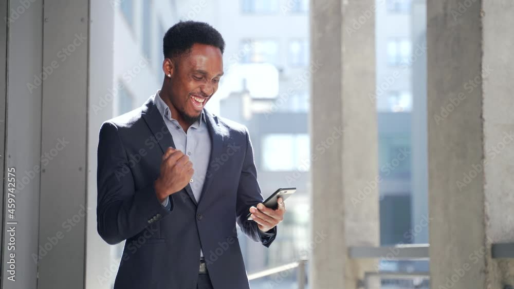 Exited African American Businessman Celebrating Achievement looking at Smartphone. Successful business man victory win with mobile phone outside. Happy Good News Success Deal, Startup Bet winning rate Exited African American Businessman Celebrating Achievement looking at Smartphone. Successful business man victory win with mobile phone outside. Happy Good News Success Deal, Startup Bet winning rate