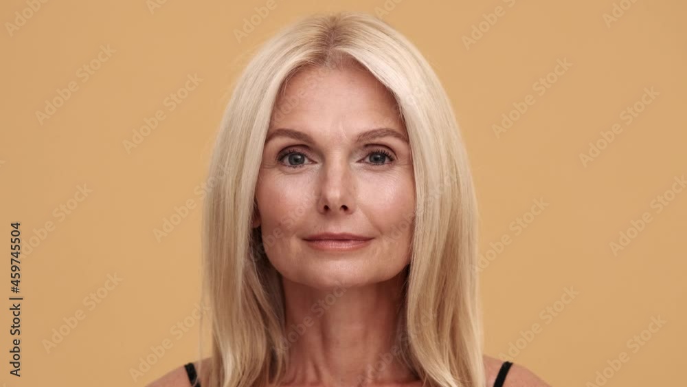 A close-up view of the smiling middle aged beautiful woman looking at the camera in the beige studio A close-up view of the smiling middle aged beautiful woman looking at the camera in the beige studio