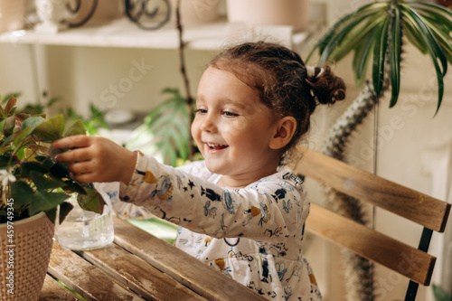 Happy mixed-race dark-haired little girl is spraying houseplants with water from a glass sprayer at home.Home gardening.Hobby concept.Biophilia design and urban jungle concept.