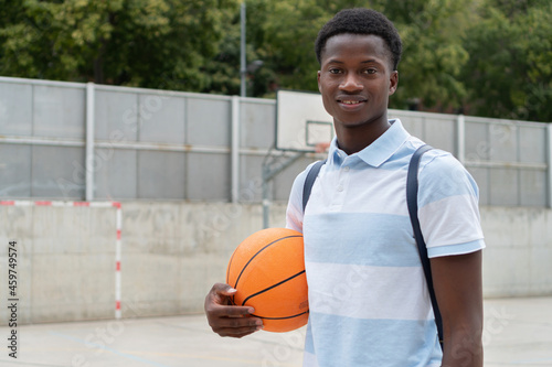 African American student back to school. Portrait of a Black teenager boy outside high school with a basketball ball in the football court.