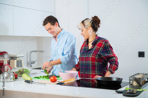 pareja joven sonriente cocinan verduras como pimientos verdes, tomate, jitomate y calabacin, con una sarten de hierro en una cocina blanca con placa de inducción