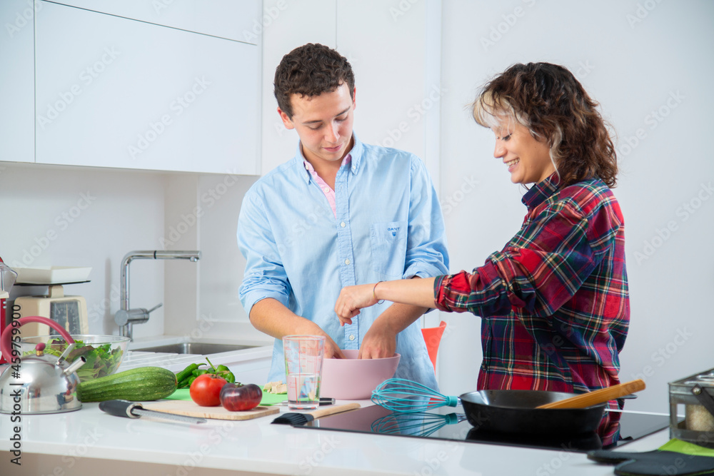 pareja joven sonriente cocinan verduras como pimientos verdes, tomate, jitomate y calabacin, con una sarten de hierro en una cocina blanca con placa de inducción