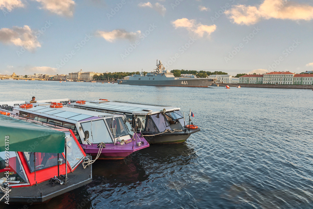 view of tourist boats and a warship on the Neva River in St. Petersburg against the blue sky and the city