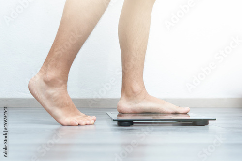 Young Latino male, climbing on a scale, gray floor, glass scale, white wall, beam of light, detail only feet, unrecognizable person, brown skin.