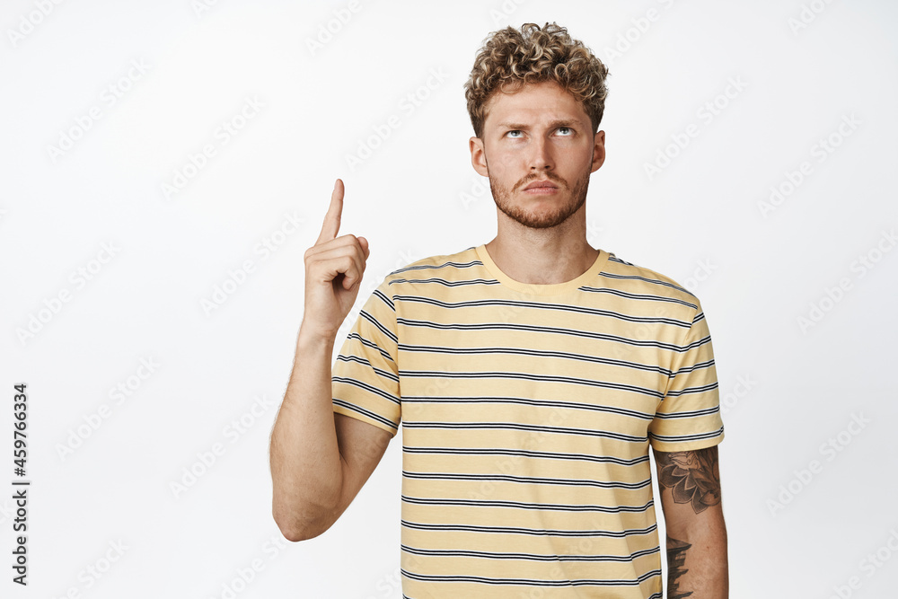 Angry and moody blond man pointing finger up, looking with upset frowning face at advertisement, standing annoyed against white background Angry and moody blond man pointing finger up, looking with upset frowning face at advertisement, standing annoyed against white background