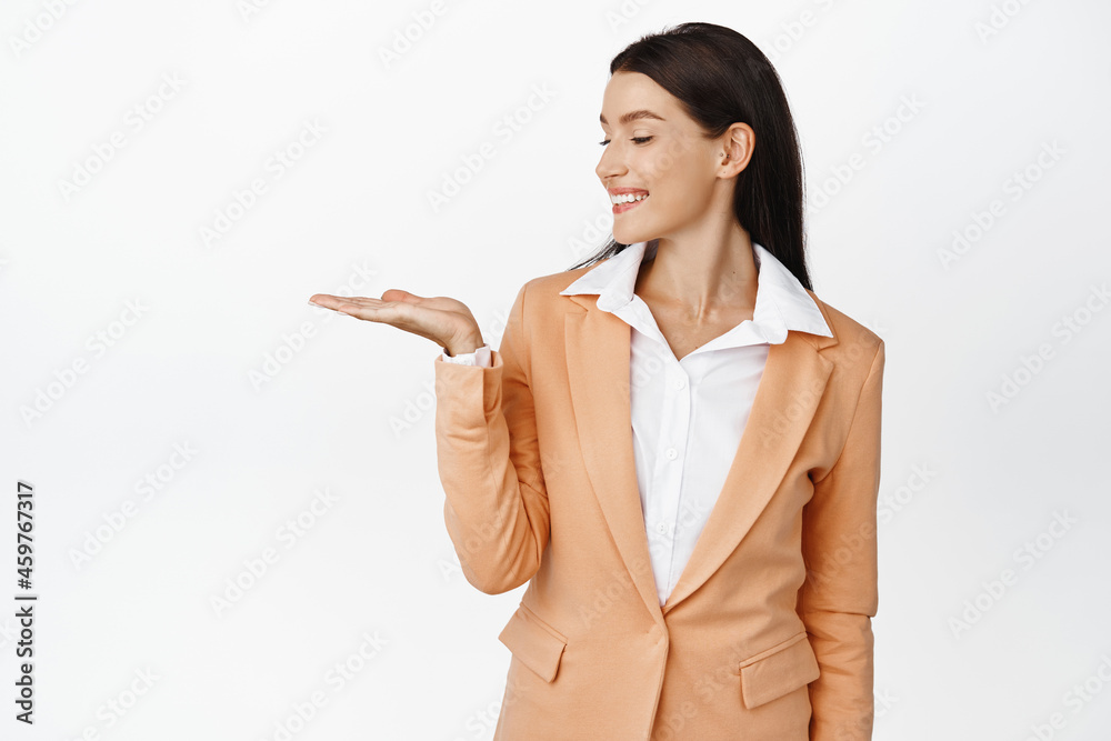 Successful corporate woman showing object on open palm, looking at her hand and smiling pleased, standing in suit against white background Successful corporate woman showing object on open palm, looking at her hand and smiling pleased, standing in suit against white background