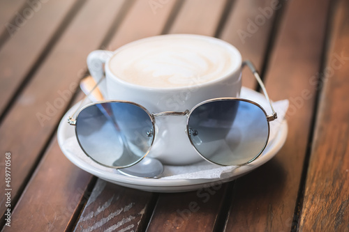 White cup of cappuccino with sunglasses stands on a wooden surface