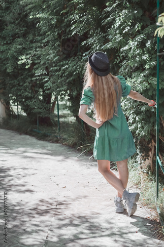 Fototapeta Naklejka Na Ścianę i Meble -  A long-haired girl in a turquoise dress and a hat stands on an alley in the park holding on to a lattice bar with her hand. Back view