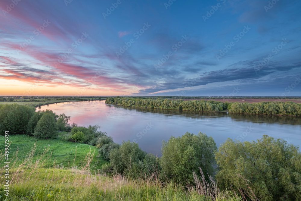 Scenic view of a beautiful summer river sunset with reflection in the water, calm water. Bright colorful cloudy sky. Lush green grass in the foreground.