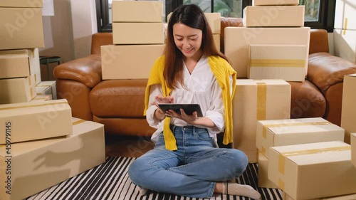 Wide shot of young Asian woman use tablet and sit among stack of carton or boxes during prepare for delivery product to customer in concept of new normal lifestyle and online selling.