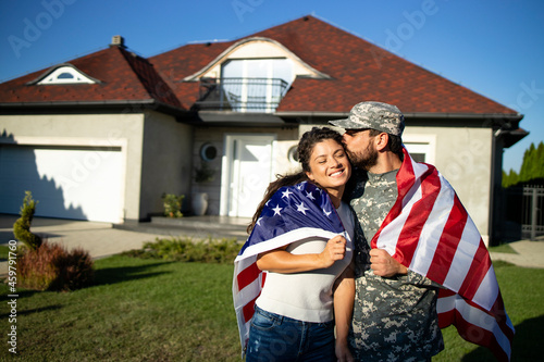 Canvas Print Portrait of husband soldier in uniform on military leave kissing his lovely wife and holding American flag in front of their house
