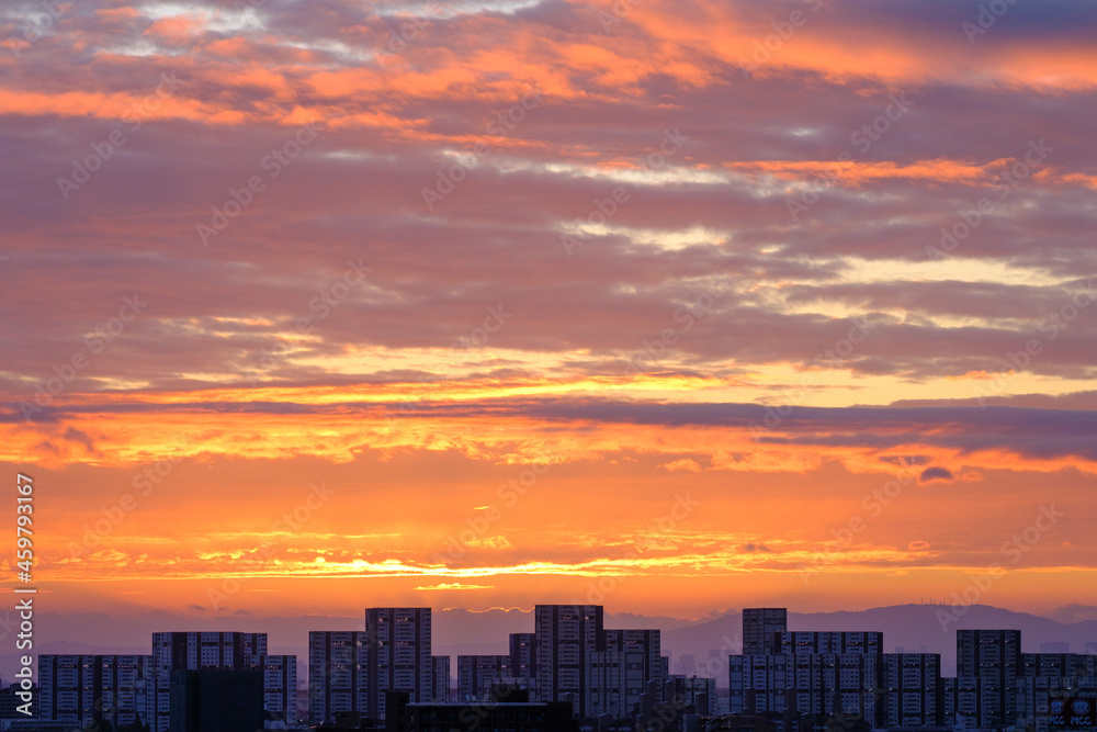 都市の夜明け、早朝ビルの隙間から太陽が昇り辺りはオレンジ色に染まる。ビルはシルエットに浮かぶ。