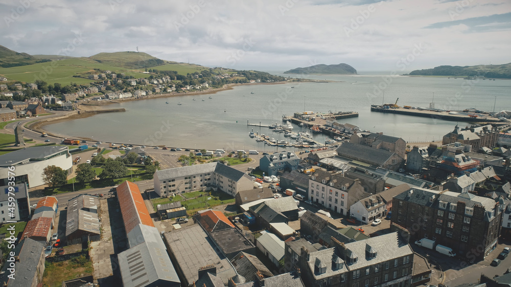 Fototapeta premium Harbor town cityscape aerial. Seascape with yachts, ships at urban street with road. Cars drive at traffic highway. Modern buildings at ocean shore of Campbeltown, Scotland, Europe rise-up drone shot