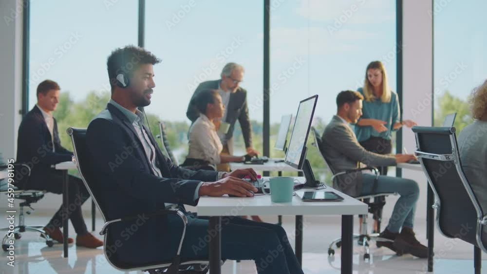 Working in modestly furnished room with large windows. Mixed race businessman using headset for communication. Modern open space office interior. Working in modestly furnished room with large windows. Mixed race businessman using headset for communication. Modern open space office interior.