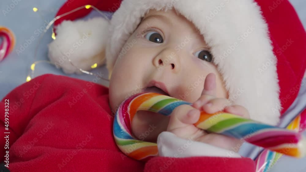 Smiling funny newborn baby in santa claus hat, red bodysuit play on white soft bed lick sweet. Concept holiday Christmas, candys , happy new year, infants, childhood. Close-up, Smiling funny newborn baby in santa claus hat, red bodysuit play on white soft bed lick sweet. Concept holiday Christmas, candys , happy new year, infants, childhood. Close-up,